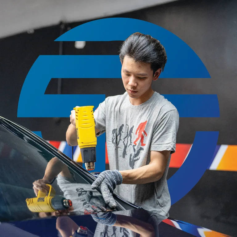 Technician using a heat gun on a car windshield, set against TGA's oversized blue logomark